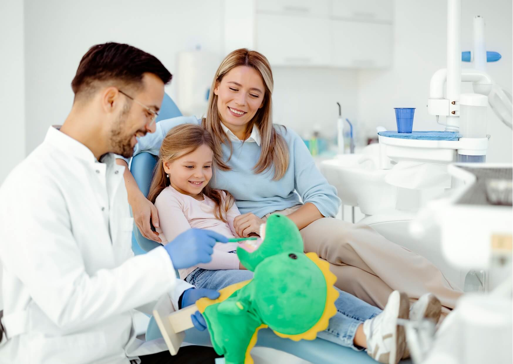 Pediatric dentist using a stuffed dinosaur to demonstrate brushing techniques and prepare a child for a comfortable first dental visit with a parent present.