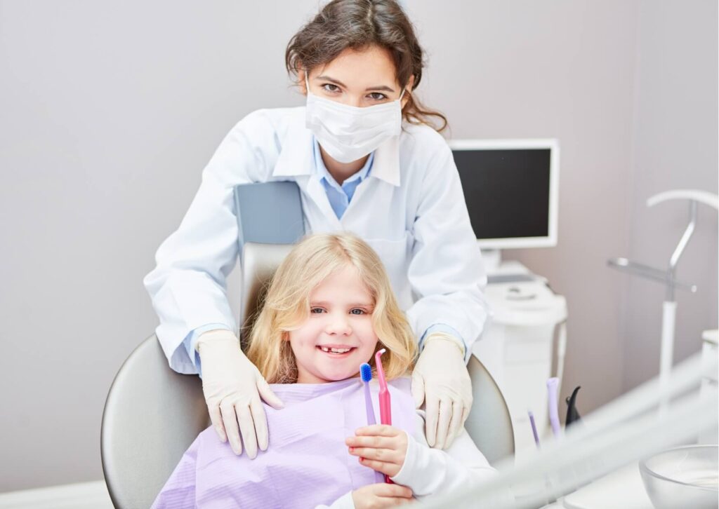 Young child smiling in a dental chair while holding a toothbrush as a pediatric dentist helps prepare the child for a positive dental visit.
